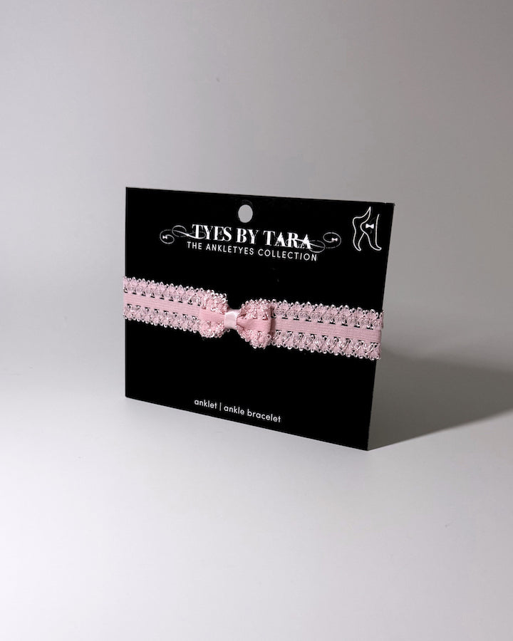 A pink elastic bow anklet with bow centered on the anklet on a black retail card on a softly lit background