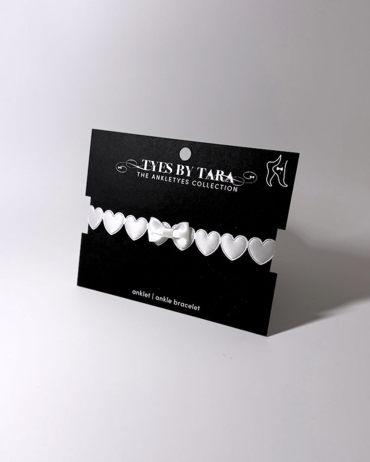 A white anklet bracelet with a row of hearts and a central bow, displayed on a black branded card labeled 'TYES BY TARA THE ANKLETS COLLECTION'.