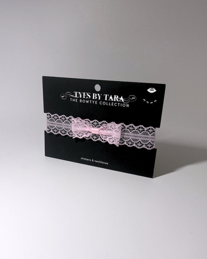 A light pink lace garter bow choker with a silver-tone chain extender with clasp, laid flat on a light gray background displayed on black TYES BY TARA branded black card