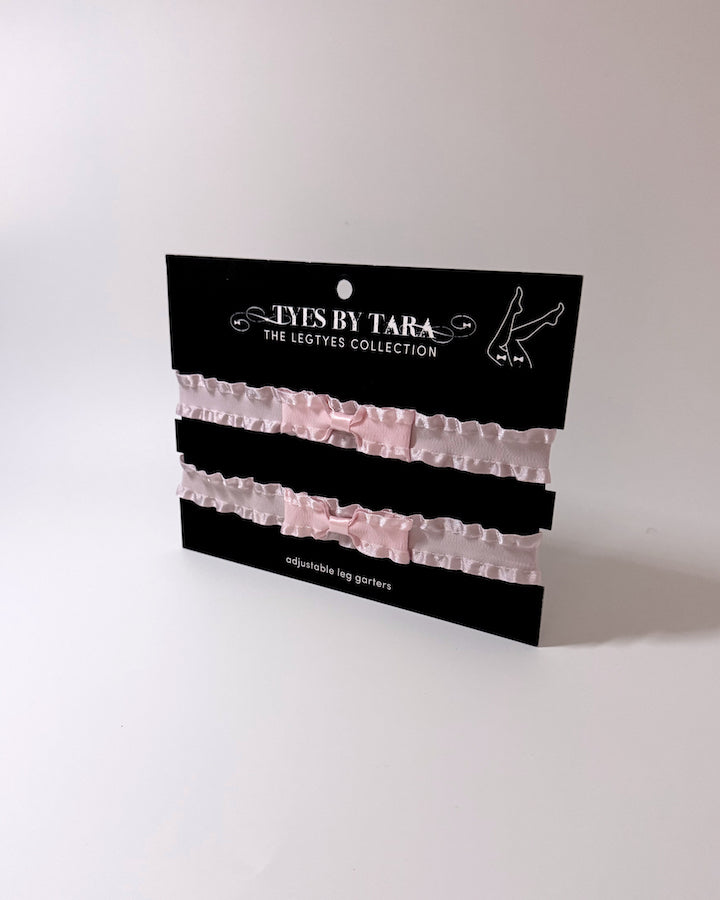 Two pale pink ruffled leg garters with bows, displayed on a black card labeled 'EYES BY TARA THE LEGTIES COLLECTION' against a plain light background.