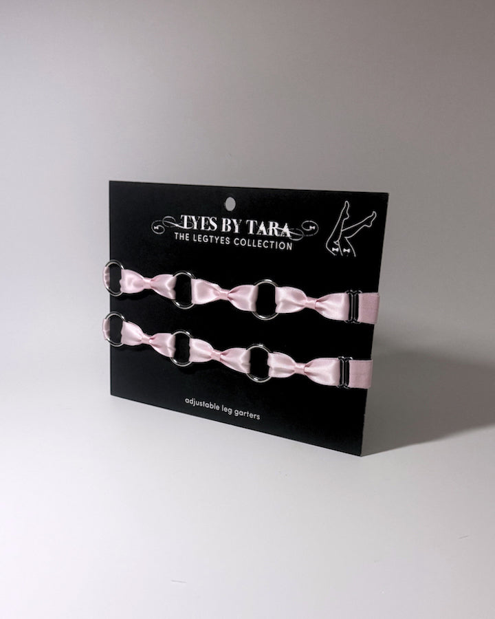 Two light pink satin bow leg garters with silver metal rings, displayed on a black card labeled 'TYES BY TARA THE LEGTYES COLLECTION'.