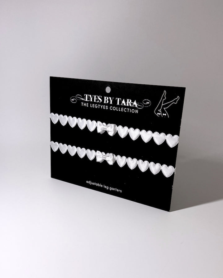 A black display card for 'Tyes By Tara The Legtyes Collection' holding two rows of white heart-shaped leg garters, photographed on a light gray background.