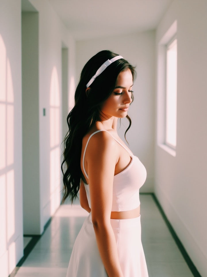Woman wearing satin bow headband in a light pink top and white skirt standing in a sunlit room.