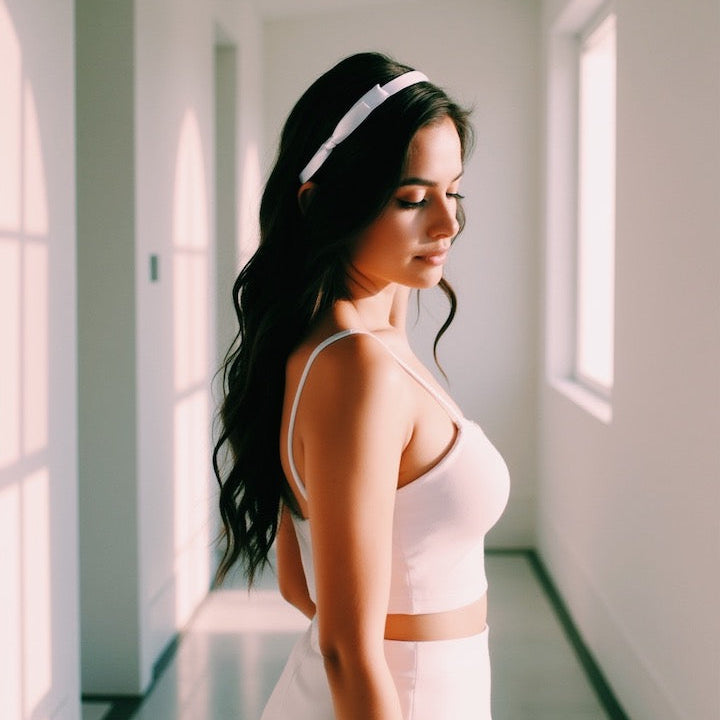 Woman wearing satin bow headband in a light pink top and white skirt standing in a sunlit room.