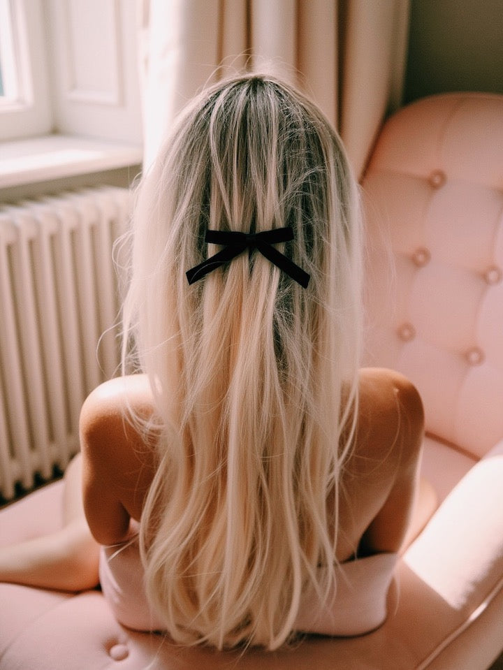Model with long blonde hair wearing a black bow hair bow sitting on a pink couch.
