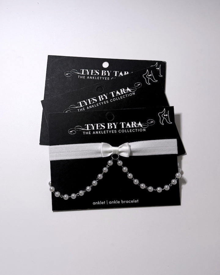 A pearl anklet anklet with a white bow and pearl chain, displayed on black retail cards. The brand 'TYES BY TARA' is clearly printed on the packaging.