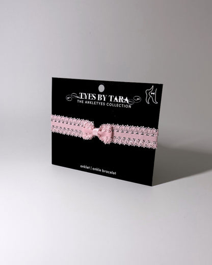 A pink elastic bow anklet with bow centered on the anklet on a black retail card on a softly lit background 