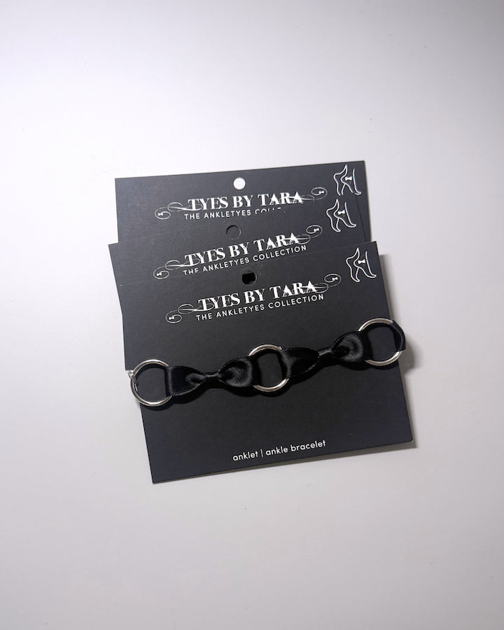 A black anklet with silver metal o rings, displayed on a black branded card that reads 'TYES BY TARA THE ANKLET COLLECTION'.