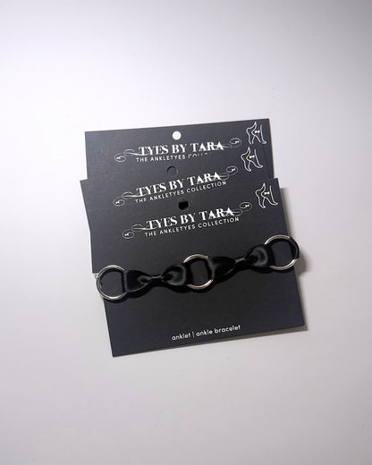 A black anklet with silver metal o rings, displayed on a black branded card that reads 'TYES BY TARA THE ANKLET COLLECTION'.