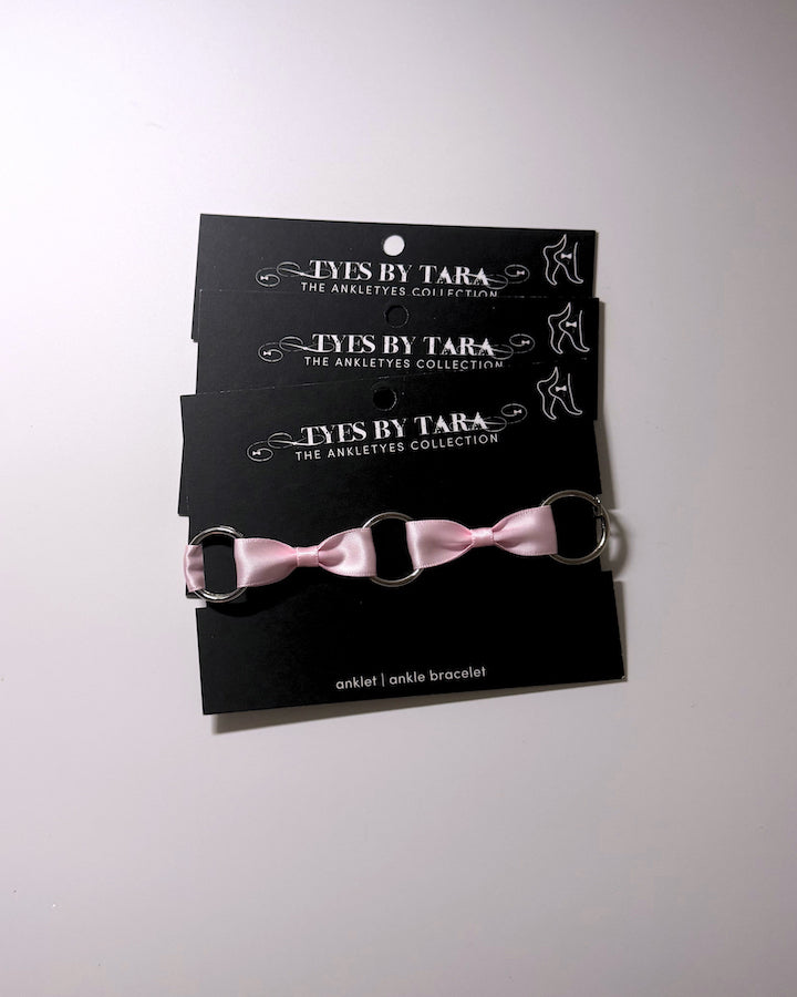 Three black display cards stacked vertically, each holding a matching pink ribbon anklet with silver metal rings. The cards are branded 'TYES BY TARA THE ANKLETYES COLLECTION'.