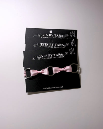 Three black display cards stacked vertically, each holding a matching pink ribbon anklet with silver metal rings. The cards are branded 'TYES BY TARA THE ANKLETYES COLLECTION'.