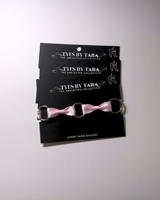 Three black display cards stacked vertically, each holding a matching pink ribbon anklet with silver metal rings. The cards are branded 'TYES BY TARA THE ANKLETYES COLLECTION'.
