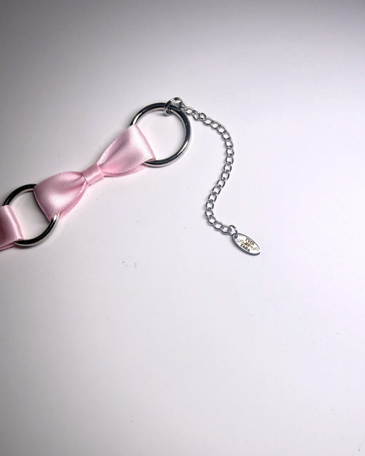 A close-up of a pink satin bow accessory attached to black metal O-rings and a silver chain with a small dangling charm, photographed on a plain light background.