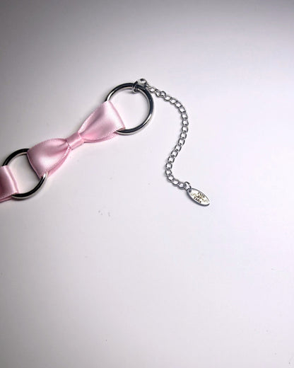 A close-up of a pink satin bow accessory attached to black metal O-rings and a silver chain with a small dangling charm, photographed on a plain light background.