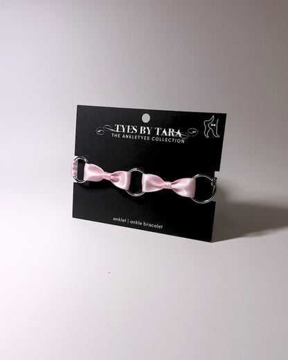 A pink satin bow anklet with silver metal rings, displayed on a black branded card that reads 'TYES BY TARA THE ANKLETS COLLECTION'.