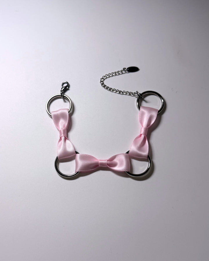 A pink satin bow anklet with silver metal rings and a silver chain extender, laid flat on a light gray background.