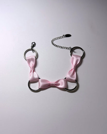 A pink satin bow anklet with silver metal rings and a silver chain extender, laid flat on a light gray background.