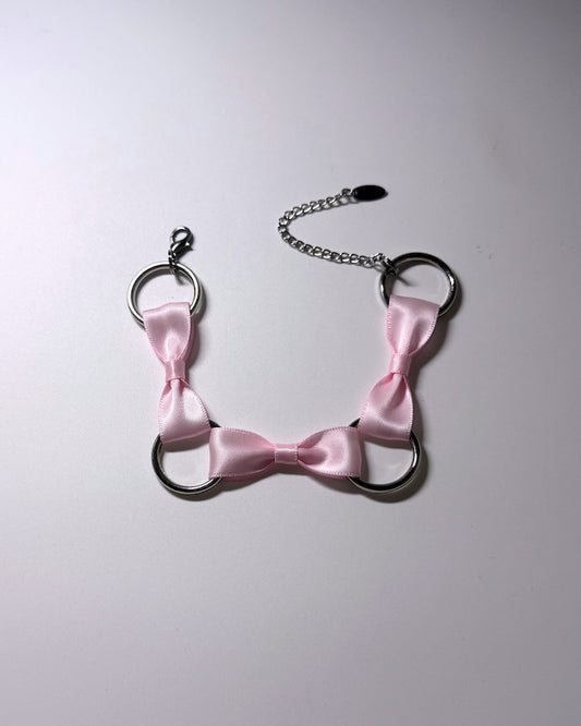 A pink satin bow anklet with silver metal rings and a silver chain extender, laid flat on a light gray background.