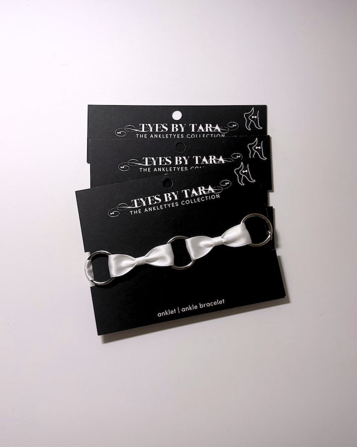 White bow anklet bracelets in a 3 pack displayed on a black branded card labeled 'TYES BY TARA'.
