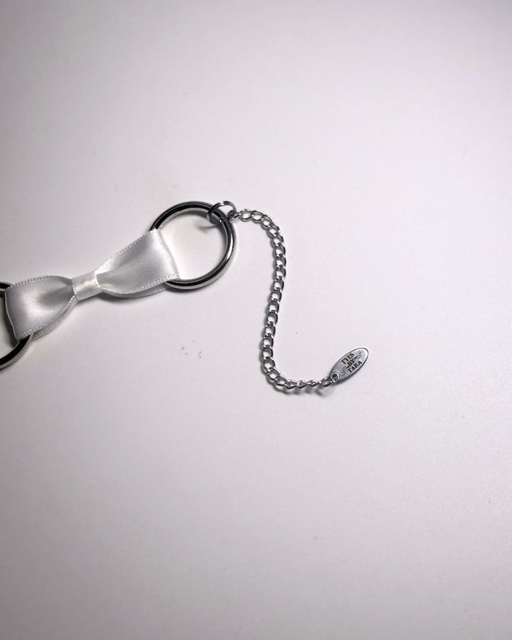 A close-up of a silver-toned metal extender chain and oval tyes by tara logo tag on a plain light background.