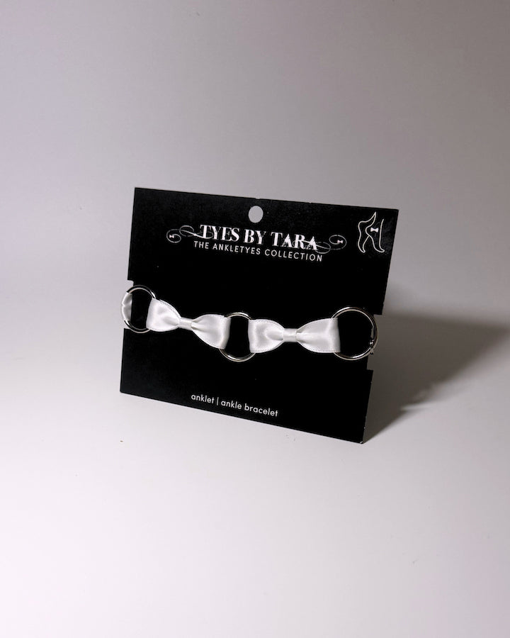 A white satin bow anklet with silver metal rings, displayed on a black branded card that reads 'TYES BY TARA THE ANKLETS COLLECTION'.