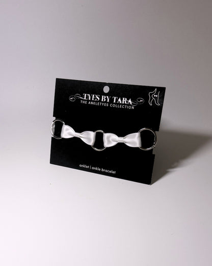 A white satin bow anklet with silver metal rings, displayed on a black branded card that reads 'TYES BY TARA THE ANKLETS COLLECTION'.