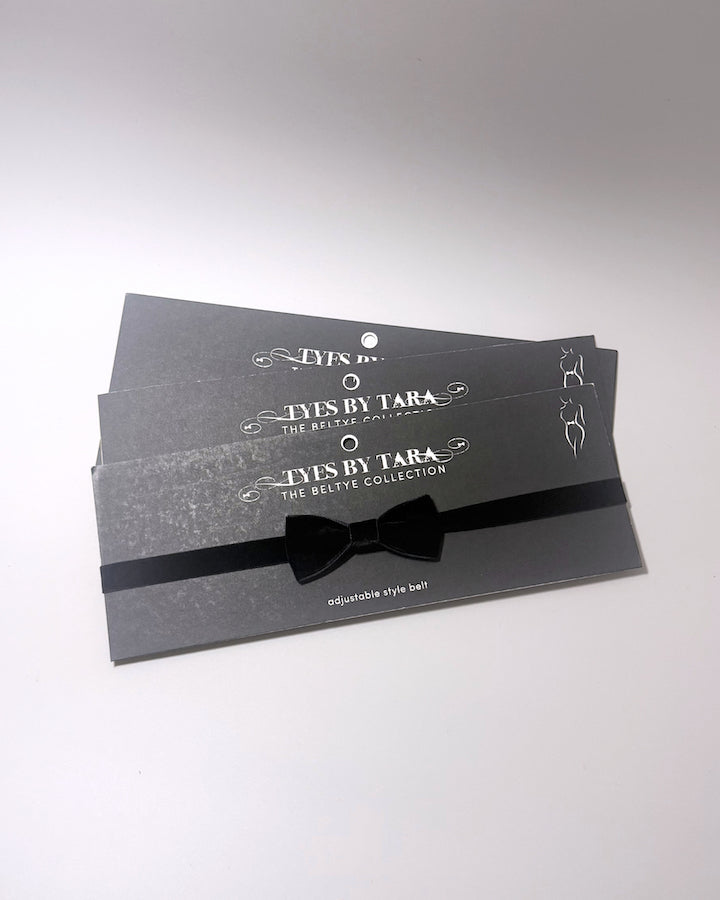 Three black adjustable velvet bow belts from Tyes By Tara, neatly stacked on their branded dark gray packaging cards with white text.