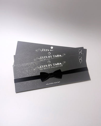 Three black adjustable velvet bow belts from Tyes By Tara, neatly stacked on their branded dark gray packaging cards with white text.