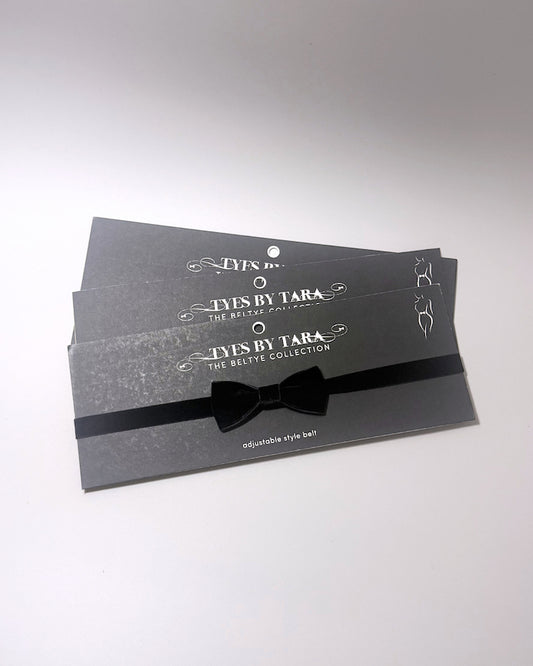 Three black adjustable velvet bow belts from Tyes By Tara, neatly stacked on their branded dark gray packaging cards with white text.
