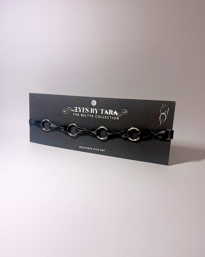 A black bow belt with silver o rings on a black retail card