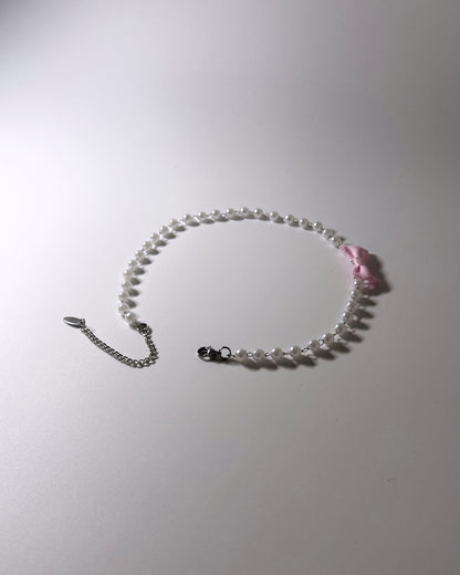 A delicate beaded necklace with small white pearls, a silver chain extender, and a small pink ribbon bow accent, displayed on a light gray background.