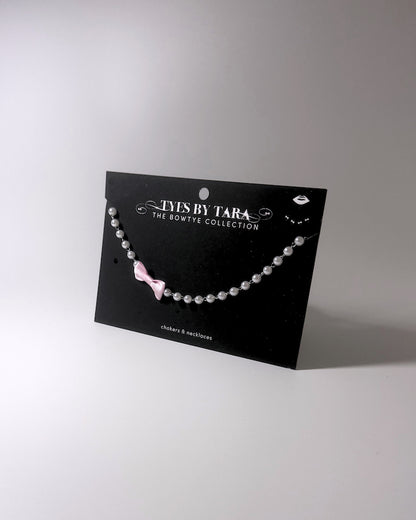 A pearl beaded necklace with a small pink bow charm, displayed on a black card from 'TYES BY TARA THE BOWTYE COLLECTION' against a neutral gray background.