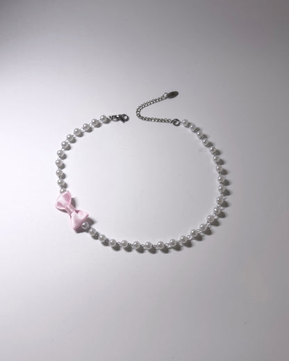 A delicate necklace made of small white pearls with a small pink satin bow accent and a silver chain extender clasp, shown on a plain light gray background.