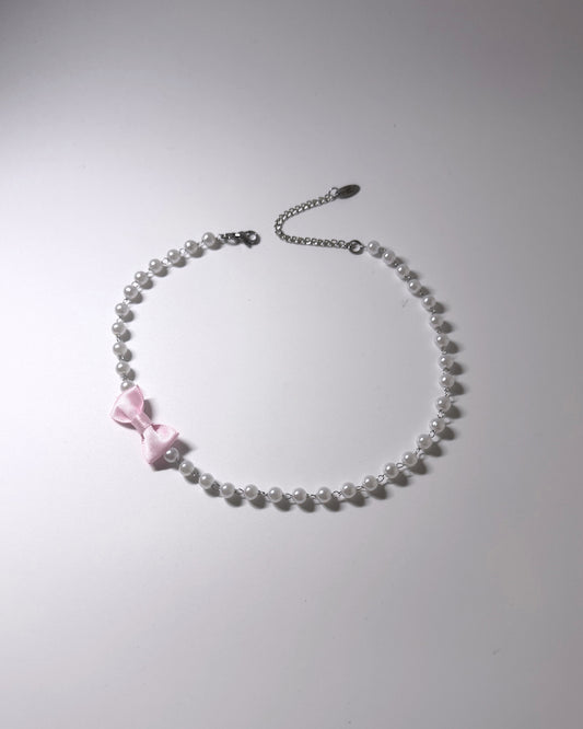 A delicate necklace made of small white pearls with a small pink satin bow accent and a silver chain extender clasp, shown on a plain light gray background.
