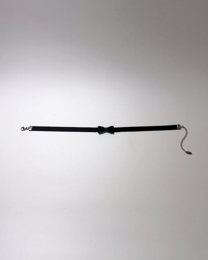 A black choker collar featuring a central bow tie detail, silver-tone metal hardware on both ends, and a short dangling chain with a small tag, displayed on a light gray background.
