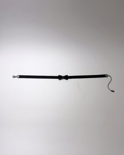 A black choker collar featuring a central bow tie detail, silver-tone metal hardware on both ends, and a short dangling chain with a small tag, displayed on a light gray background.