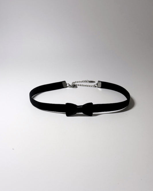 A black velvet choker collar featuring a central black bow accent and a silver ball chain extender clasp, displayed on a neutral light gray background.