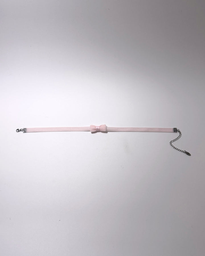 A pale pink velvet choker collar featuring a central bow detail, silver metal hardware, and an adjustable chain extender, displayed against a neutral light gray background.