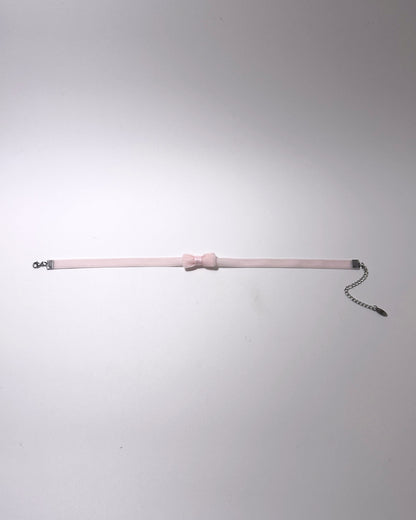 A pale pink velvet choker collar featuring a central bow detail, silver metal hardware, and an adjustable chain extender, displayed against a neutral light gray background.
