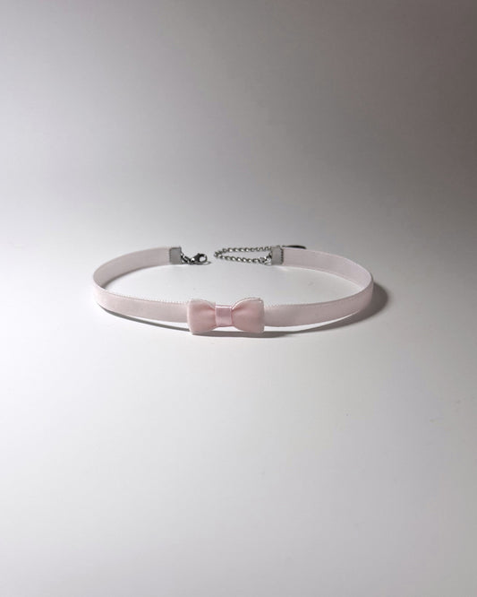 A light pink velvet choker collar featuring a matching bow at the center and a silver metal clasp with a short chain extender, displayed on a neutral gray background.