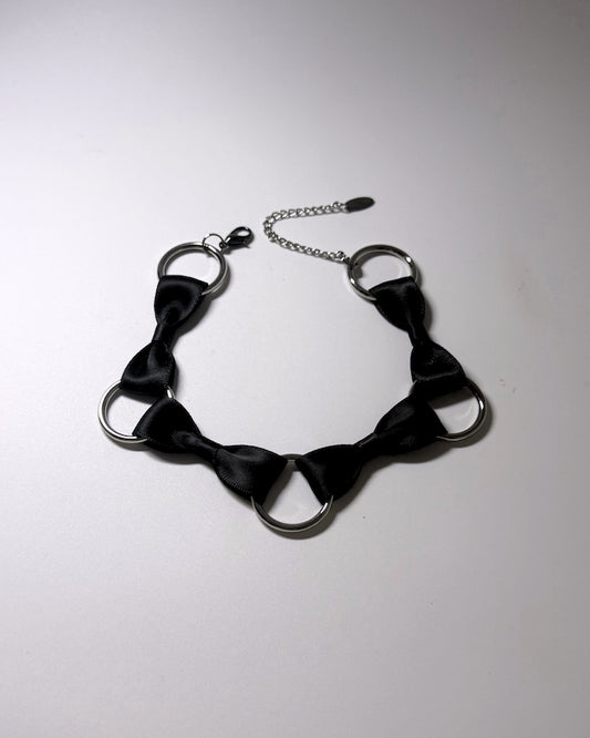 A black satin bow tie collar with silver-tone metal rings and a silver chain extender, displayed on a light gray background.