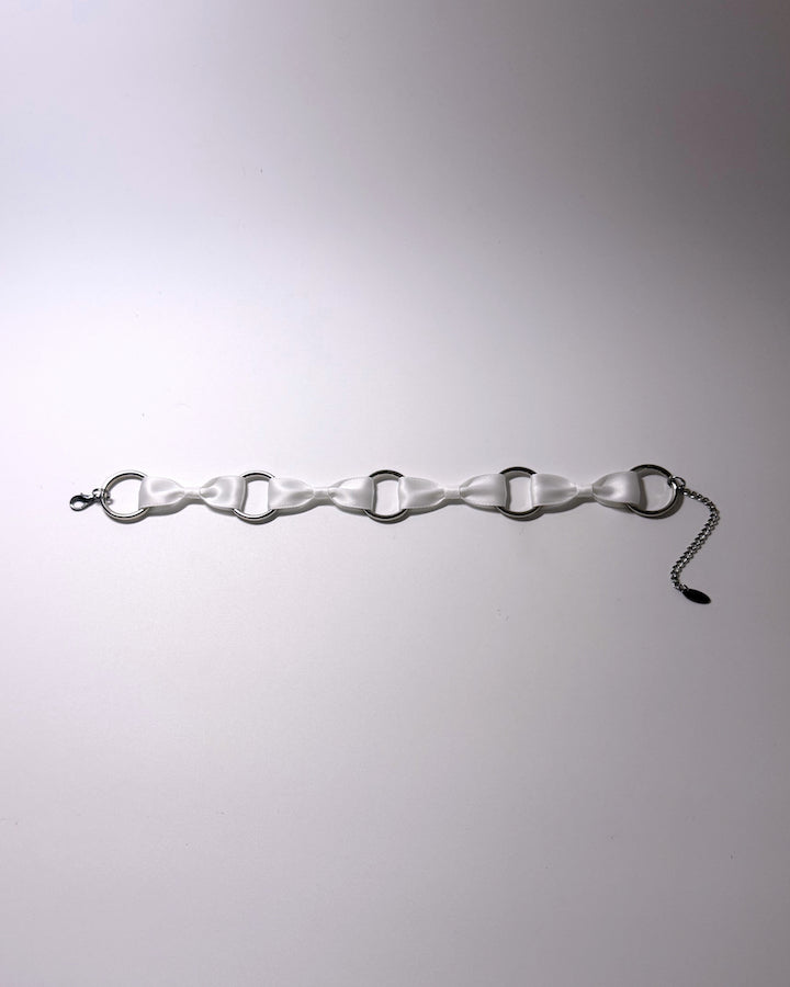 A white bow choker with silver-toned metal O-rings and a silver chain extender, laid flat on a light gray background.