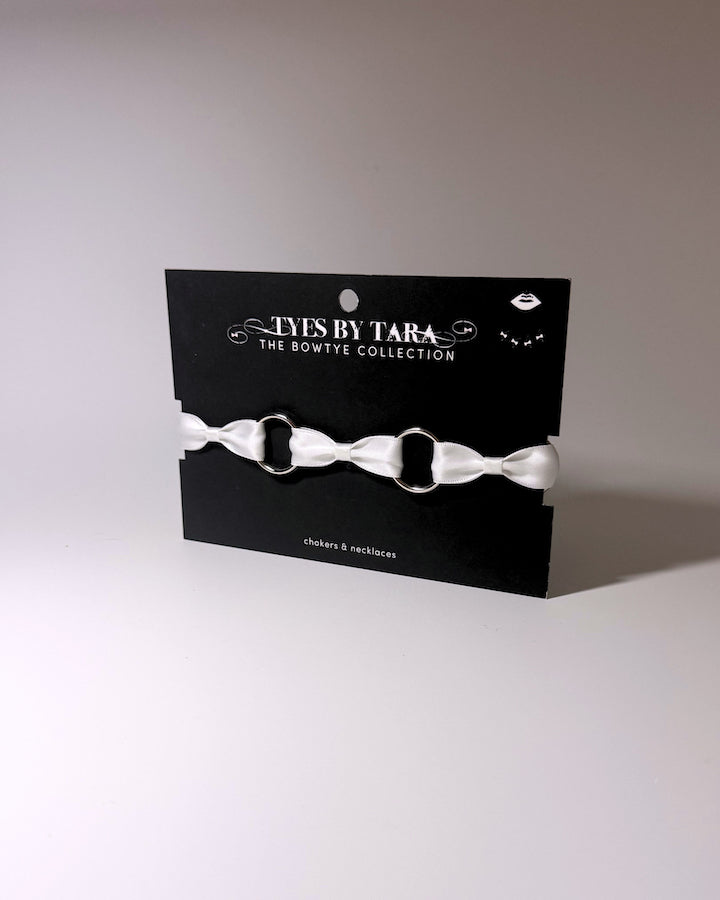 A white satin bow choker displayed on a black branded card from 'Tys Is By Tara The Bowtye Collection'.