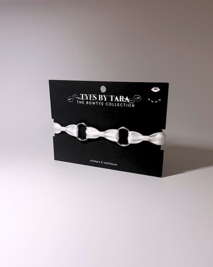 A white satin bow choker displayed on a black branded card from 'Tys Is By Tara The Bowtye Collection'.