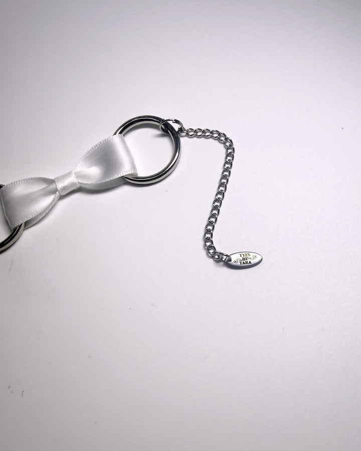 A close-up of a silver metal chain extender and a small engraved TYES BY TARA tag, attached to a white satin bow, on a plain light background.
