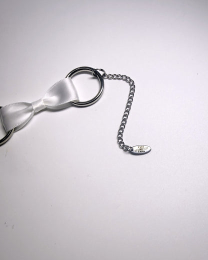 A close-up of a silver metal chain extender and a small engraved TYES BY TARA tag, attached to a white satin bow, on a plain light background.