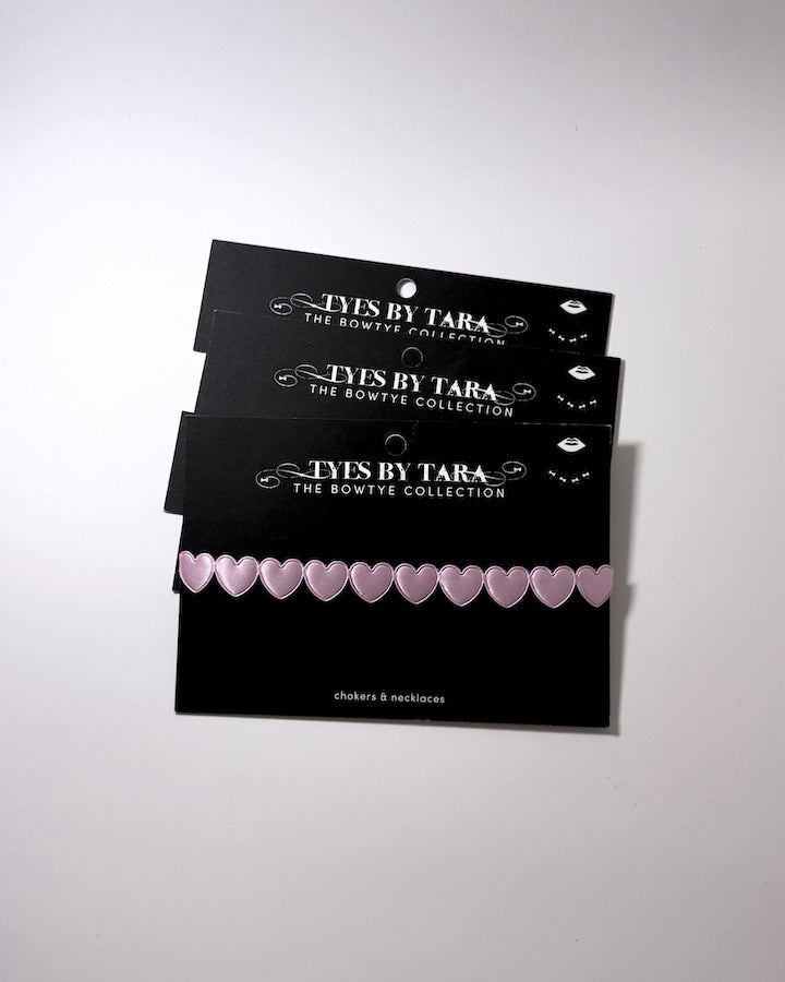 Three black display cards stacked on a light gray background, each holding a row of small pale pink heart-shaped bows. The cards are branded 'TYES BY TARA THE BOWTYE COLLECTION'.