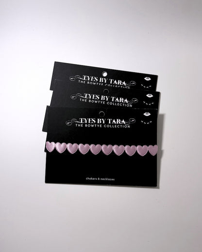 Three black display cards stacked on a light gray background, each holding a row of small pale pink heart-shaped bows. The cards are branded 'TYES BY TARA THE BOWTYE COLLECTION'.