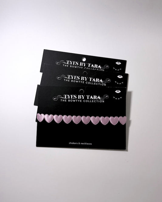 Three black display cards stacked on a light gray background, each holding a row of small pale pink heart-shaped bows. The cards are branded 'TYES BY TARA THE BOWTYE COLLECTION'.