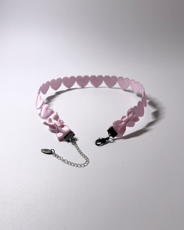 A light pink heart-patterned choker collar with a silver lobster clasp and an adjustable silver chain extender, displayed on a neutral light gray background.